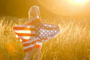 skön ung flicka innehav ett amerikan flagga i de vind i en fält av råg. sommar landskap mot de blå himmel. horisontell orientering. foto