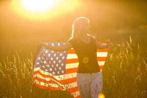 ung Lycklig flicka löpning och Hoppar sorglös med öppen vapen över vete fält. innehav USA flagga. tonad bild. selektiv fokus. foto