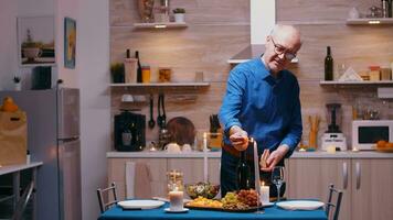 senior man belysning de ljus väntar hans fru för en romantisk middag. äldre gammal Make framställning festlig måltid med hälsosam mat för årsdag firande, Sammanträde nära de tabell i kök. foto