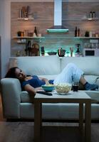 utmattad kvinna tittar på TV visa liggande på soffa i levande rum. trött ensam olycklig ung lady i pijamas avkopplande på bekväm soffa i främre av tv känsla uttråkad äter snacks sent på natt foto