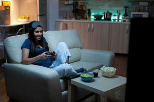 fast besluten kvinna spelar video spel i levande rum på natt. upphetsad gamer kvinna Sammanträde på soffa, spelar och vinnande video spel använder sig av trösta och trådlös kontroller. foto