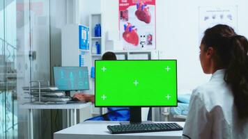 läkare använder sig av dator med grön skärm attrapp i sjukhus och assistent promenader ut av skåp bär blå enhetlig. läkare i vit täcka arbetssätt på övervaka med krom nyckel i klinik skåp till kolla upp patient diagnostisera. foto