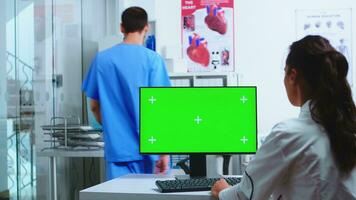 läkare använder sig av dator med krom nyckel och assistent innehav patient röntgen i sjukhus skåp. skrivbordet med utbytbar grön skärm i medicinsk klinik medan läkare är kontroll patient radiografi för diagnostisera. foto