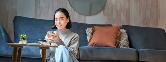 teknologi och människor. ung eleganta asiatisk kvinna sitter på Hem med henne smartphone, textning meddelande, använder sig av Ansökan på mobil telefon foto