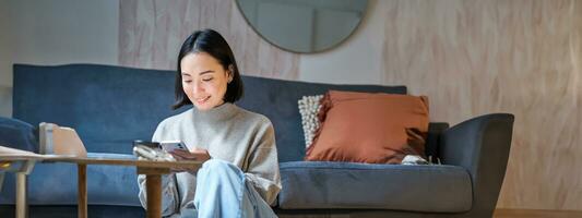 teknologi och människor. ung eleganta asiatisk kvinna sitter på Hem med henne smartphone, textning meddelande, använder sig av Ansökan på mobil telefon foto