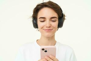 porträtt av uppriktig ung kvinna, lyssnar musik i trådlös hörlurar, innehav smartphone, tittar på videoklipp på mobil telefon, stående över vit bakgrund foto