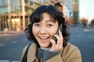 porträtt av Lycklig koreanska flicka, samtal på mobil telefon, utseende överraskad och Lycklig, mottar positiv bra Nyheter över telefon konversation, står på gata av stad foto