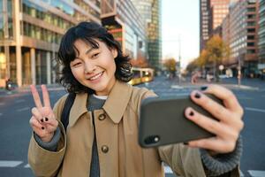 Lycklig asiatisk flicka, turist tar Foto i stad Centrum, visar fred tecken på smartphone kamera, gör selfie under sightseeing med leende glad ansikte