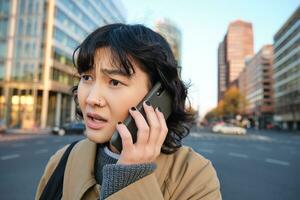 stänga upp porträtt av bekymrad asiatisk kvinna, samtal på mobil telefon och hör dålig Nyheter, utseende orolig, känner frustrerad förbi telefon konversation, står på gata foto