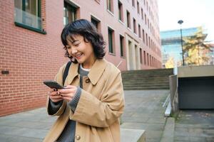eleganta koreanska hipster flicka, promenader i stad Centrum, skickar text meddelande, användningar plats Karta Ansökan på smartphone medan gående ner de gata, bär dike täcka foto