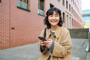 eleganta koreanska hipster flicka, promenader i stad Centrum, skickar text meddelande, användningar plats Karta Ansökan på smartphone medan gående ner de gata, bär dike täcka foto