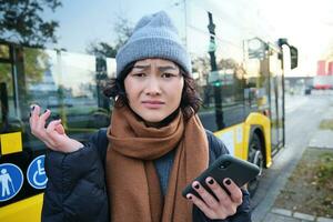 porträtt av förvirrad asiatisk flicka, stående på buss sluta, innehav mobil telefon, ser chockade och upprörd, besviken förbi offentlig transport fördröjning foto