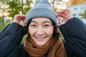 porträtt av söt asiatisk flicka i hatt och scarf, promenader runt om stad i kyligt vår väder, ler och utseende Lycklig, sitter i parkera nära grön gräs foto