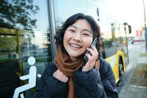 cellulär teknologi och människor begrepp. eleganta asiatisk flicka samtal på mobil telefon, gör en telefon ringa upp, står nära buss sluta och har konversation foto