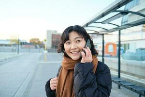 skön leende koreanska flicka, väntar på buss sluta, använder sig av offentlig transport, talande på mobil telefon, gående någonstans i stad foto