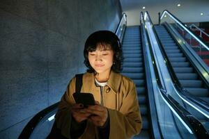 söt ung kvinna gående ner de rulltrappa till de rör, använder sig av tunnelbana metro till pendla till arbete eller universitet, stående med smartphone foto