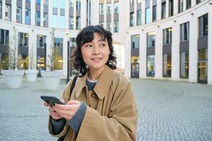 eleganta koreanska flicka i hörlurar, lyssnar musik och användningar mobil telefon, står i stad Centrum, väntar för någon på gata och skrivningar text meddelande på henne smartphone foto