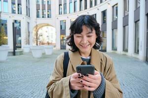 eleganta koreanska flicka i hörlurar, lyssnar musik och användningar mobil telefon, står i stad Centrum, väntar för någon på gata och skrivningar text meddelande på henne smartphone foto
