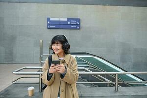 porträtt av eleganta asiatisk hipster flicka, lyssnar musik i hörlurar, utseende på henne telefon, turist ser på Karta för en sätt till sightseeing, dricka kaffe till gå foto