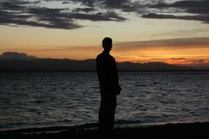 silhuett av en ung man stående förbi de sjö njuter de solnedgång. fredlig atmosfär i natur foto