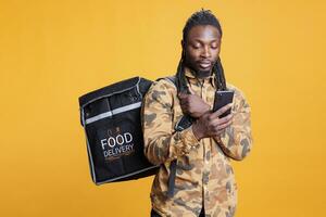 afrikansk amerikan kurir spårning kund adress på smartphone innan leverera beställa under lunch tid. deliveryman innehav termisk ryggsäck full med hämtmat mat över gul bakgrund foto