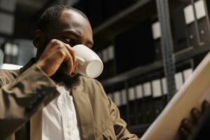 privat forskare dricka kaffe medan granskning polis dokumentation. afrikansk amerikan man detektiv- arbetssätt sent och läsning brottslighet materia fil i byrå kontor på natt tid foto