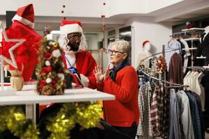 anställd i kostym presenter kläder till kvinna, sökande för mode objekt som presenterar. man porträtterar santa claus hälsning kunder och ger bistånd under Semester försäljning. foto