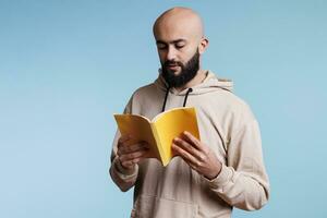 ung omtänksam fokuserade arab man läsning ny från bok med gul omslag. koncentrerad skallig skäggig person innehav öppen mjukt täcke lärobok och studerar med fundersam uttryck foto