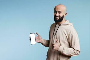 leende arab man reklam app på pekskärm smartphone, visning tom falsk upp. glad person som visar mobil telefon med tömma vit skärm och ser på kamera med tumme upp gest foto