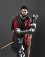 medeltida riddare på grå bakgrund. porträtt av brutal smutsig ansikte krigare med kedjepost rustning röda och svarta kläder och stridsyxa foto