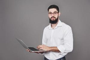 stilig affärsman i glasögon och kostym som håller bärbar dator i händerna och skriver något. sidovy. isolerad grå bakgrund foto