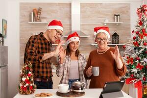 Lycklig familj bär santa hatt hälsning avlägsen föräldrar fira jul säsong tillsammans under uppkopplad videosamtal möte konferens i xmas dekorerad kök. barn njuter vinter- Semester foto