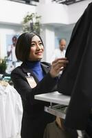 Kläder Lagra leende asiatisk anställd fastsättning jacka knappar medan bär mannekäng i formell kläder. handla köpcenter boutique kvinna arbetstagare organisering damkläder för försäljning foto