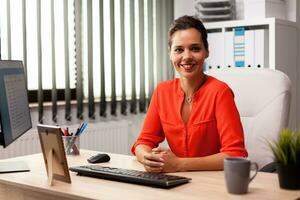 verkställande frilansare finansiera företagare bär röd blus leende på kamera i arbetsplats. framgångsrik självsäker kvinna i marknadsföring Sammanträde på skrivbord i arbetsplats använder sig av dator. foto