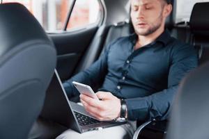 en ung affärsman som arbetar på bärbar dator och pratar i telefon medan han sitter i bilens rygg. arbetar i rörelse, uppskattar sin tid foto
