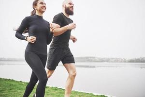 par joggar och springer utomhus i parken nära vattnet. ung skäggig man och kvinna som tränar tillsammans på morgonen foto