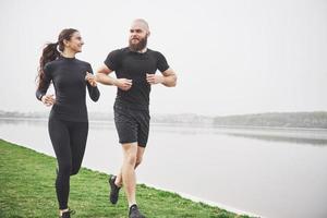 par joggar och springer utomhus i parken nära vattnet. ung skäggig man och kvinna som tränar tillsammans på morgonen foto