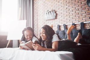 två vackra afrikanska flickor i sängkläder leende sitter på sängen hemma vaknade på morgonen på en solig dag.