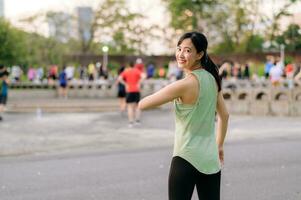 kvinna joggare. passa ung asiatisk kvinna med grön sportkläder aerobics dansa övning i parkera och njuter en friska utomhus. kondition löpare flicka i offentlig parkera. wellness varelse begrepp foto