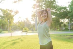 kvinna joggare. passa ung asiatisk kvinna med grön sportkläder stretching muskel i parkera innan löpning och njuter en friska utomhus. kondition löpare flicka i offentlig parkera. wellness varelse begrepp foto