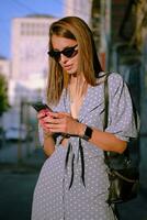 blond flicka i lång blå klänning, solglasögon och en små svart handväska är använder sig av henne smartphone medan gående ensam i de stad. närbild. foto