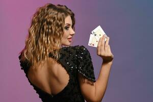 brunhårig lady i svart skinande klänning som visar två spelar kort, Framställ på färgrik bakgrund. spelande, poker, kasino. närbild, tillbaka se. foto