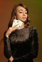 brunett kvinna i svart sammet klänning och Smycken som visar två ess, Framställ mot färgrik studio bakgrund. spelande, poker, kasino. närbild. foto