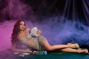 flicka i gyllene klänning spelar poker på kasino, innehav kontanter, om på tabell med pommes frites, kort på Det. svart, rök bakgrund. spelande. närbild. foto