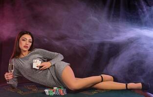 flicka i grå klänning spelar poker på kasino, innehav kort, glas av champagne, om på tabell med pommes frites på Det. svart, rök bakgrund. närbild. foto