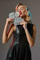 blond flicka i svart eleganta klänning innehav och kissing några pengar, Framställ mot grå bakgrund. hasardspel underhållning, poker, kasino. närbild. foto