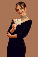brunett modell, bar axlar, i svart klänning och Smycken. leende, som visar två spelar kort, Framställ på brun bakgrund. poker, kasino. närbild foto