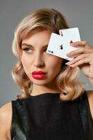 blond flicka i svart läder klänning som visar två spelar kort, Framställ mot grå bakgrund. hasardspel underhållning, poker, kasino. närbild. foto