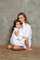 inomhus- porträtt av en skön mor med henne charmig liten dotter Framställ mot en grå vägg. foto