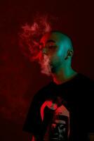 närbild studio skott av en ung skallig kille vaping, blåser ut en moln av rök på röd bakgrund. foto